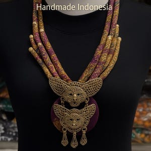 Op de afbeelding: Een luxe batik ketting met een Kutubaru broche. De ketting heeft een ontwerp met meerdere strengen met goud en paarse stof. De broche is een gedetailleerd goudmetalen ontwerp met een cirkelvormige paarse achterkant. De tekst luidt "Luxury Batik Necklace" en "Handmade Indonesia."