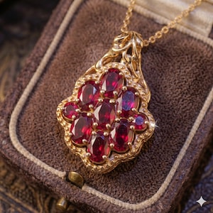 May include: A gold pendant necklace featuring a cluster of oval-cut, deep red gemstones. The pendant has an ornate, filigree design and is set in a gold frame. The necklace is displayed in a brown jewelry box.