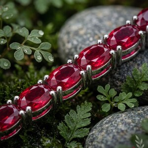 May include: A silver bracelet featuring oval-cut, vibrant red gemstones. The bracelet is set against a backdrop of green foliage, moss, and small stones, creating a natural and elegant display. The gemstones are the focal point, showcasing their rich color and brilliance.