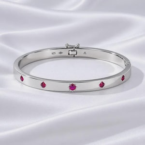 May include: A silver-toned hinged bangle bracelet featuring a central star-shaped setting with a round red stone, flanked by four smaller square-cut red stones. The interior band is engraved with the text 925 and a small triangular hallmark symbol.