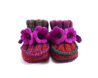 May include: A pair of handmade baby booties. The booties are knit with a combination of red, purple, and brown yarn. Each bootie features a large, bright purple flower on the front. The booties are designed to keep a baby's feet warm and cozy.