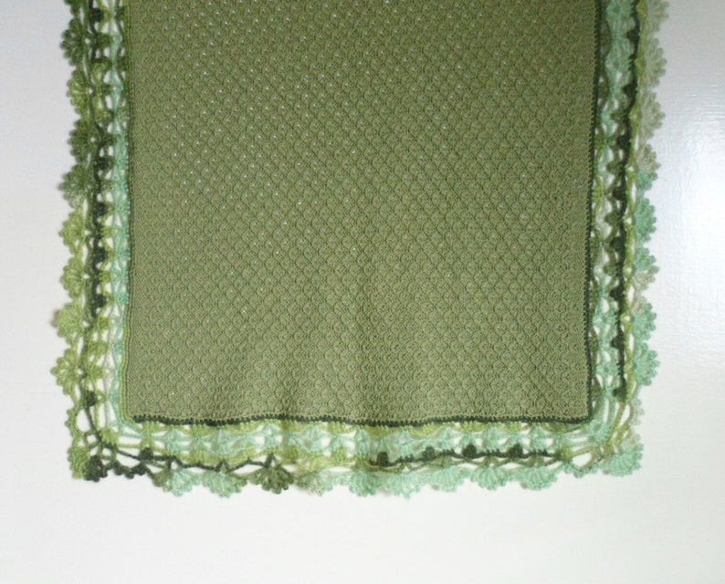 May include: A green crocheted blanket with a delicate lace border. The blanket is made with a textured stitch and has a subtle ombre effect in the border.