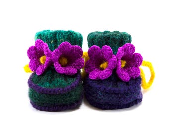May include: A pair of hand-knitted booties in dark green and purple yarn, featuring two vibrant purple crocheted flowers with yellow centers on the front of each bootie and bright yellow drawstring ties at the back of the ankle cuffs.