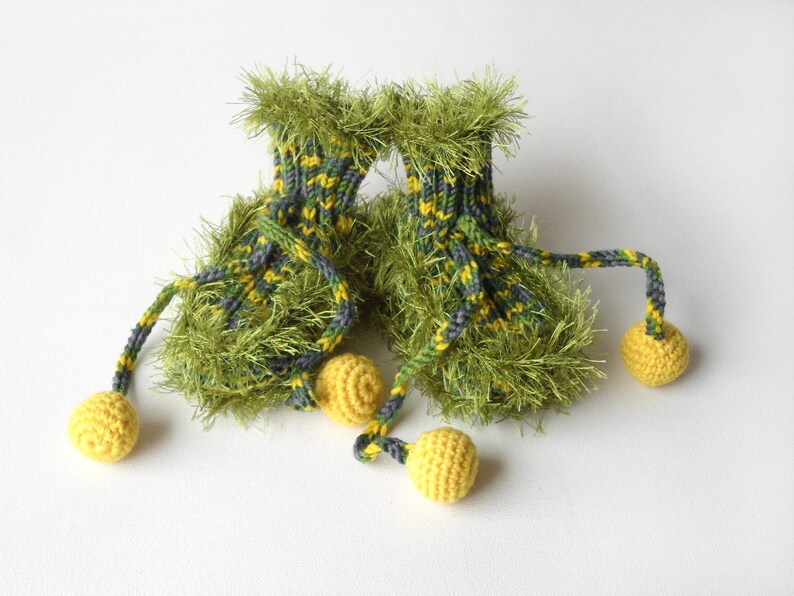 May include: A pair of green and yellow knitted baby booties with fuzzy trim and yellow pom-poms attached to the laces.
