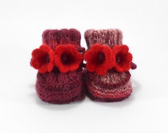 May include: A pair of handmade baby booties in shades of burgundy and pink, adorned with bright red flower accents. The knitted booties are displayed on a white background, showcasing the intricate details of the yarn and floral embellishments.