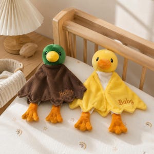 Pode incluir: Dois paninhos de boca de pato de pelúcia com corpos de tecido macio e pés bordados. Um pato tem cabeça verde-escura e corpo castanho, o outro cabeça amarela e corpo amarelo. Ambos apresentam a palavra Baby bordada em castanho.