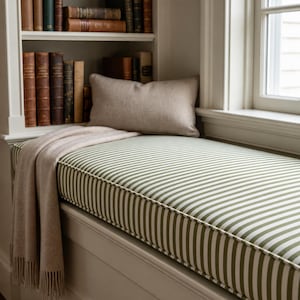 May include: A window seat cushion featuring a green and white vertical striped pattern, accompanied by a solid beige rectangular throw pillow and a draped beige throw blanket with fringe, positioned near a bookshelf with vintage books.