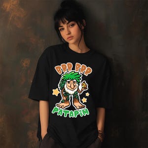 Pu&ograve; includere: T-shirt nera con un personaggio dei cartoni animati con capelli verdi e foglie, che tiene in mano un uccellino. Il testo "BRP BRP PATAPIM" &egrave; visualizzato in un font giocoso, con stelle. La maglietta ha una vestibilit&agrave; ampia.