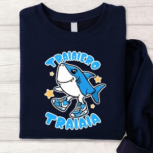 Pu&ograve; includere: Felpa blu navy con uno squalo dei cartoni animati con scarpe da ginnastica blu. Lo squalo sorride e ha le parole "TRAIAIERO TRAIAIA" in un carattere a bolle blu. Sono inclusi anche accenti di stelle gialle.
