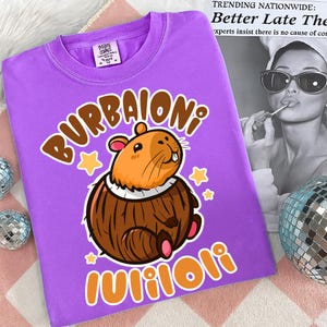 Pu&ograve; includere: Una maglietta viola con un disegno di capibara dei cartoni animati e le parole "BURBAIONI" e "Wiiloli". Il capibara &egrave; marrone e arancione, con stelle gialle. La maglietta ha un girocollo.