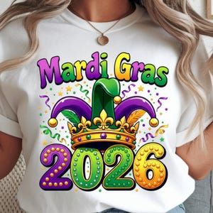May include: White t-shirt featuring a colorful Mardi Gras design. The graphic includes the words "Mardi Gras" in purple and yellow, a green and purple jester hat, a gold crown, and the numbers "2026" in purple, green, and yellow.