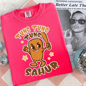 Pu&ograve; includere: Una maglietta rosa acceso con un personaggio dei cartoni animati che tiene una mazza da baseball, con le parole "Tung Tung Tung Sahur" in marrone. La maglietta ha un'etichetta di "The Home Company".