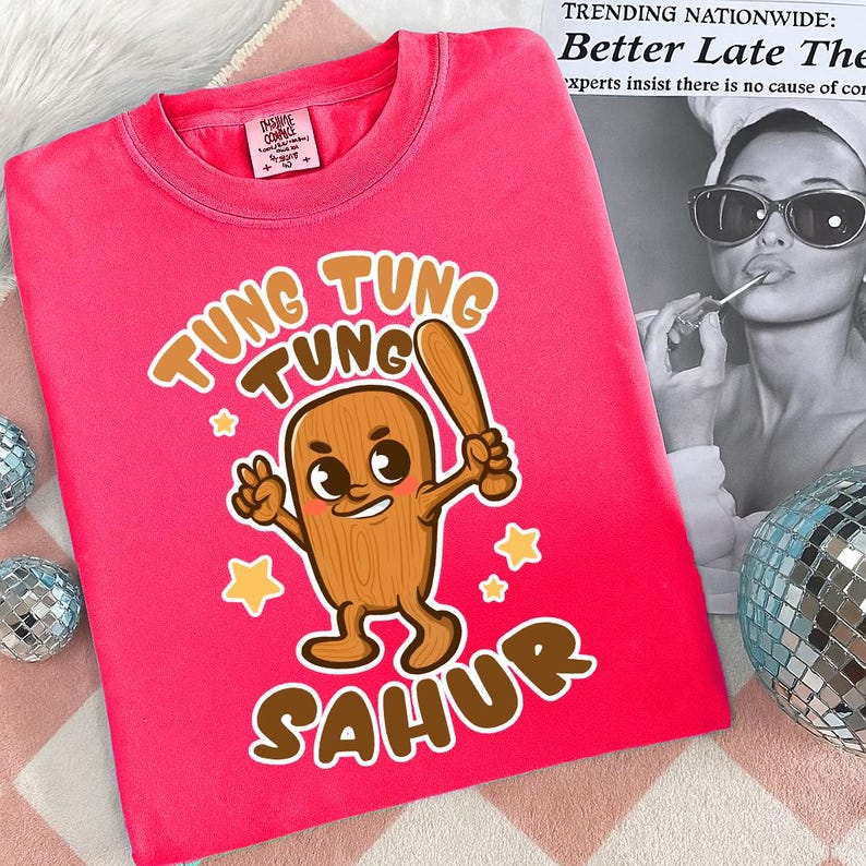 Pu&ograve; includere: Una maglietta rosa acceso con un personaggio dei cartoni animati che tiene una mazza da baseball, con le parole "Tung Tung Tung Sahur" in marrone. La maglietta ha un'etichetta di "The Home Company".