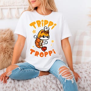 Pu&ograve; includere: T-shirt bianca con un design di gamberetti dei cartoni animati con accenti arancioni e gialli, con occhiali, bolle e stelle. La maglietta mostra le parole "TRIPPI TROPPI" in arancione.