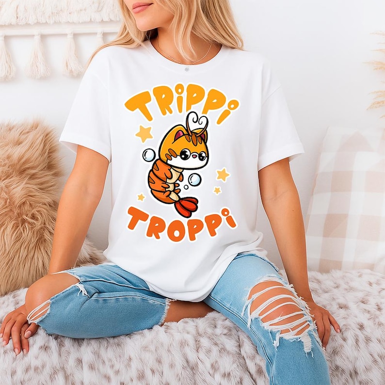 Pu&ograve; includere: T-shirt bianca con un design di gamberetti dei cartoni animati con accenti arancioni e gialli, con occhiali, bolle e stelle. La maglietta mostra le parole "TRIPPI TROPPI" in arancione.