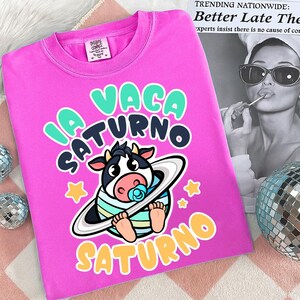 Pu&ograve; includere: Una maglietta rosa acceso con un cartone animato di una mucca che indossa un anello spaziale a righe e un ciuccio. Il testo "LA VACA SATURNO" e le stelle sono in un font giocoso. Girocollo.