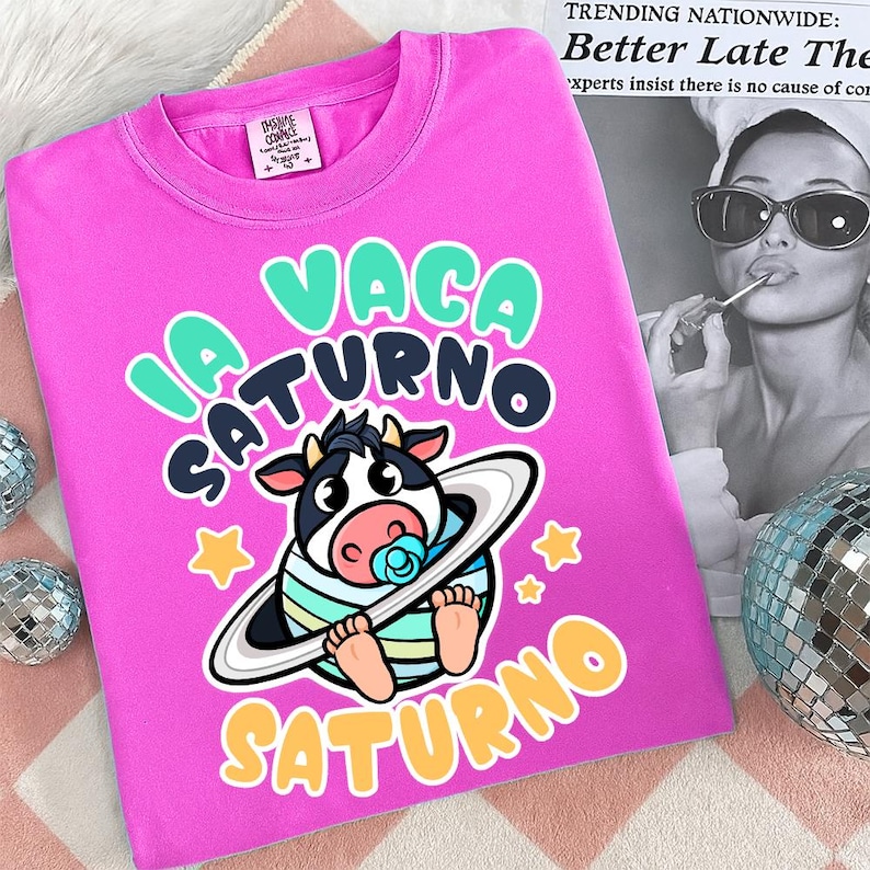 Pu&ograve; includere: Una maglietta rosa acceso con un cartone animato di una mucca che indossa un anello spaziale a righe e un ciuccio. Il testo "LA VACA SATURNO" e le stelle sono in un font giocoso. Girocollo.