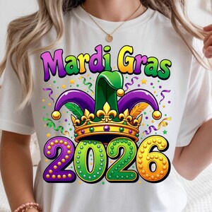 May include: White t-shirt featuring a colorful Mardi Gras design. The graphic includes the words "Mardi Gras" in purple, green, and yellow, along with a jester hat, crown, and the year "2026". Confetti and stars complete the festive look.
