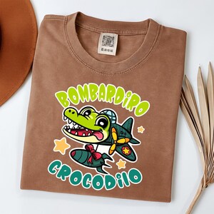 Pu&ograve; includere: T-shirt marrone con un coccodrillo dei cartoni animati con un casco da pilota, con un aereo e una bomba. Il testo recita "Bombardiro Crocodilo" in verde. Il design include stelle gialle e un fiocco rosso.