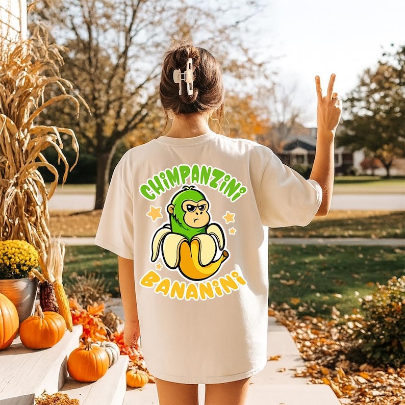 Pu&ograve; includere: T-shirt color crema con un simpatico scimpanz&eacute; dei cartoni animati che esce da una banana. Il design include le parole "CHIMPANZINI" e "BANANINI" in verde e giallo, con accenti di stelle. La maglietta fa parte di una collezione di abbigliamento.