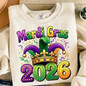 May include: Cream-colored sweatshirt with a colorful Mardi Gras graphic. The design features a jester hat, crown, and the text "Mardi Gras 2026" in purple, green, and gold. Festive confetti and stars surround the graphic.