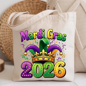 May include: Beige tote bag with the words "Mardi Gras" in purple and yellow, and the year "2026" in green and yellow. The design includes a jester hat, crown, and confetti. The bag has a simple design with two handles.