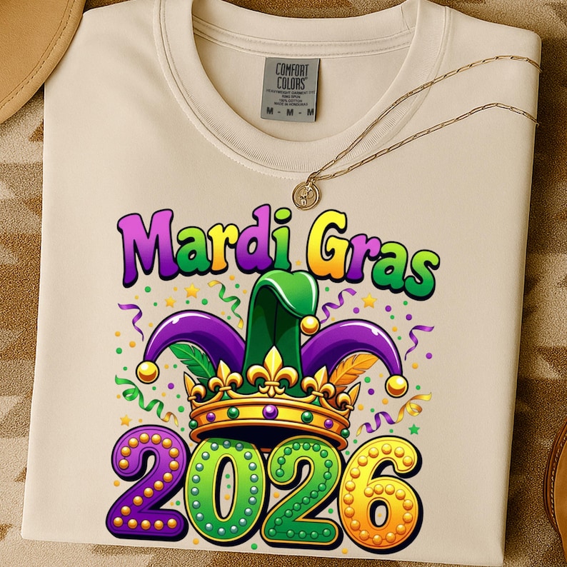 May include: Cream-colored t-shirt with a Mardi Gras design. The graphic includes the words "Mardi Gras" in purple and yellow, a jester hat, a crown, and the number "2026". A gold necklace is draped across the shirt.