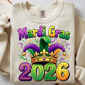 May include: Cream-colored sweatshirt with a colorful Mardi Gras design. The graphic includes the words "Mardi Gras" in purple and yellow, a jester hat, a crown, and the year "2026" in green and orange.