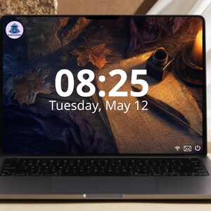 May include: A dark gray laptop with a screen displaying the time 08:25, the date Tuesday, May 12, and a background image of a quill, inkwell, and autumn leaves. A small circular logo with a robot icon is visible in the top left corner of the screen.