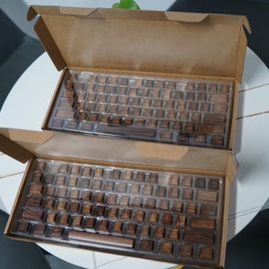 May include: Two mechanical keyboards with wooden keycaps are displayed in open cardboard boxes. The keyboards have a brown wood grain appearance and are arranged on a white table. The boxes are brown.