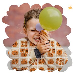 May include: A child wearing a cream jacket with a pumpkin pattern, holding a green and purple balloon. The child is smiling with their eyes closed, against a blurred pink background.