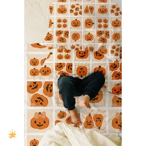 May include: A cream-coloured Halloween-themed quilt featuring a repeating pattern of orange jack-o'-lanterns with various expressions. Some pumpkins have bows. The quilt is on a bed with a person's legs and feet visible.