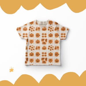 May include: A cream-coloured t-shirt with a repeating pattern of orange pumpkins, each with a different face, set within a grid. The design is ideal for Halloween, with the pumpkins in various sizes and expressions.