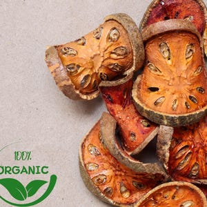 Pode incluir: Close-up de fatias de fruta bael secas, mostrando uma mistura de tons laranja e marrom avermelhado. As fatias têm uma aparência texturizada com sementes visíveis e uma estética natural e orgânica. A imagem inclui um logotipo "100% ORGANIC" verde e branco.