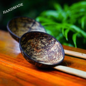 May include: Two handmade coconut shell bowls with a natural, textured pattern. The bowls are dark brown with lighter brown and tan marbling. The word "HANDMADE" is printed in white. The bowls are on a wooden surface.