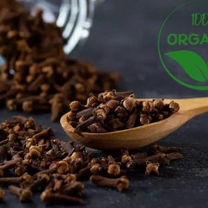 Op de afbeelding: Close-up van hele kruidnagels, sommige uit een glazen pot, andere in een houten lepel. De afbeelding bevat een groen en wit "100% ORGANIC" logo. De kruidnagels zijn donkerbruin van kleur en de achtergrond is donker.