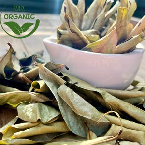 May include: Dried green and brown leaves, some curled, fill a white, curved bowl and are scattered on a wooden surface. The image includes a green and white circular graphic with the words "100% ORGANIC".
