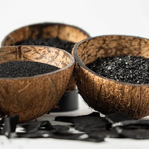 Pode incluir: Três tigelas de casca de coco castanhas preenchidas com substâncias granulares pretas de texturas variadas. As tigelas estão rodeadas por fragmentos de carvão preto espalhados. Um logótipo circular verde no canto superior direito contém o texto 100% ORGANIC.