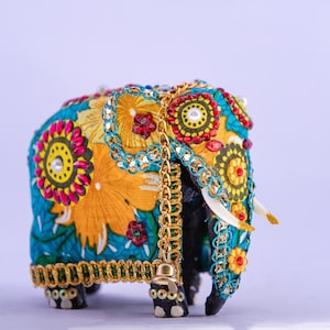 Puede incluir: Una figura decorativa de elefante, adornada intrincadamente con bordados y adornos coloridos. El elefante presenta un vibrante diseño floral turquesa y amarillo, acentuado con detalles rojos y dorados. Una cadena dorada y colmillos blancos completan el diseño.