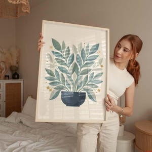 Puede incluir: Un estampado botánico enmarcado con una ilustración estilizada de una planta en una maceta azul oscuro. Las hojas son de varios tonos de azul y verde, con pequeños acentos de flores amarillas. El marco es de color madera clara.