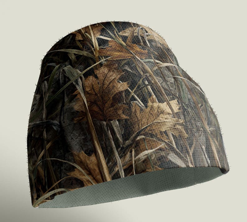 May include: A camouflage beanie hat featuring a pattern of brown leaves, green grass, and dark brown and black accents. The hat has a light green interior and is designed for outdoor activities.