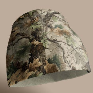 K&ouml;nnte beinhalten: Eine Camouflage-Beanie-M&uuml;tze mit einem detaillierten Waldmuster in Braun-, Gr&uuml;n- und Beiget&ouml;nen. Das Design umfasst Baumrinde, Bl&auml;tter und Tannenzapfen und erzeugt einen nat&uuml;rlichen Look. Das Innenfutter ist hellbeige.