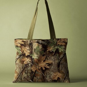 May include: A tote bag with a camouflage design, showing brown and green leaves and tree bark. It has olive green straps. The bag is rectangular and designed for carrying items.