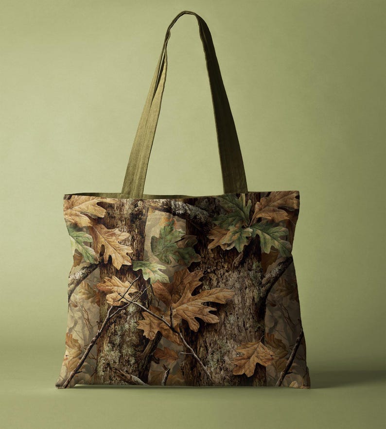 May include: A tote bag with a camouflage design, showing brown and green leaves and tree bark. It has olive green straps. The bag is rectangular and designed for carrying items.