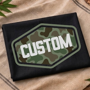 May include: A black t-shirt featuring a large, centered hexagonal emblem. The emblem has a green and brown camouflage pattern background with a thick olive green border and the word CUSTOM printed in bold, white, sans-serif capital letters.