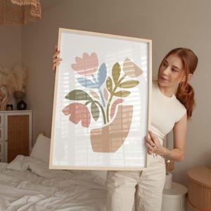May include: A framed art print featuring an abstract floral design. The artwork showcases stylized flowers in shades of pink, blue, and green, arranged in a tan vase. The frame is a light wood color, adding a natural touch to the piece.