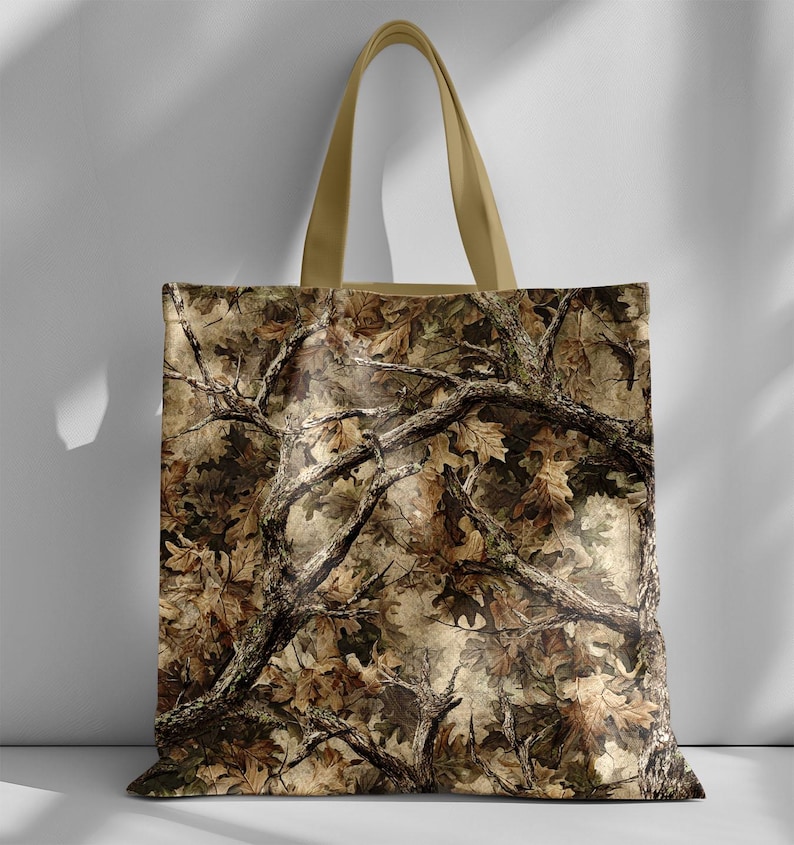 May include: A camouflage tote bag with a detailed pattern of tree branches and leaves in shades of brown, tan, and green. The bag has a beige handle and a rectangular shape, suitable for carrying various items.