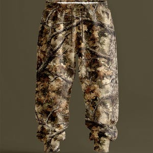 May include: Camouflage-patterned joggers with an elasticated waistband and drawstring. The trousers feature a detailed print of tree branches and leaves in shades of brown, tan, and green. The cuffs are ribbed. The overall design is intended to blend with a natural environment.