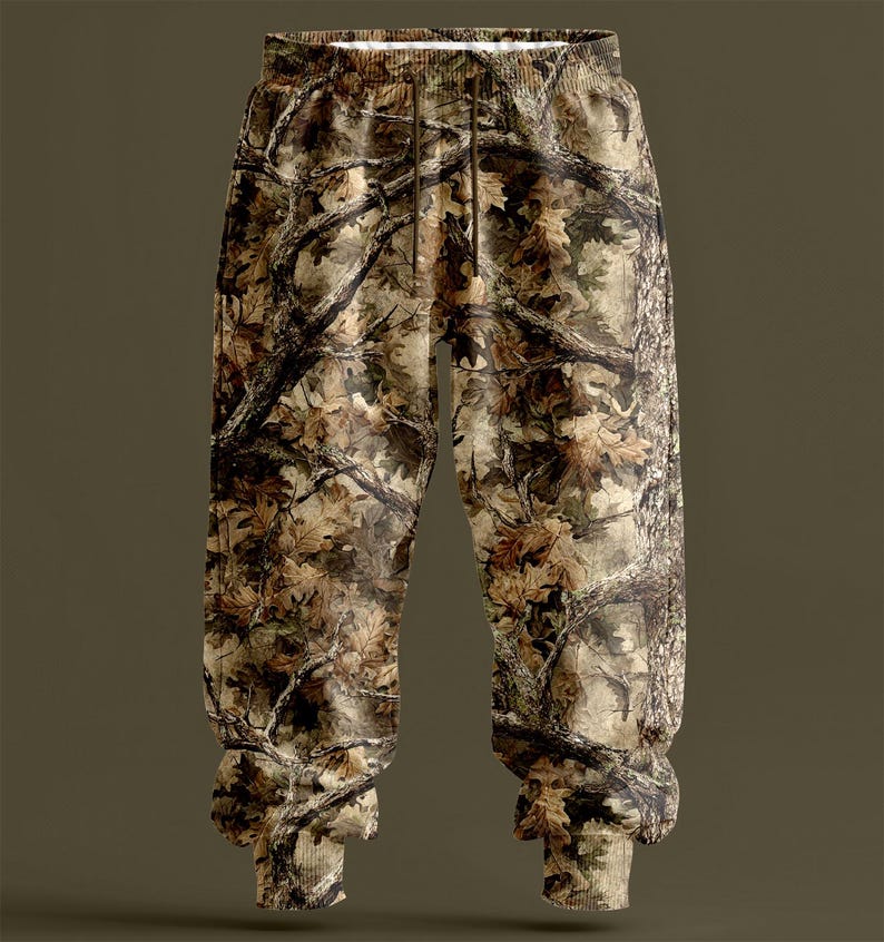 May include: Camouflage-patterned joggers with an elasticated waistband and drawstring. The trousers feature a detailed print of tree branches and leaves in shades of brown, tan, and green. The cuffs are ribbed. The overall design is intended to blend with a natural environment.