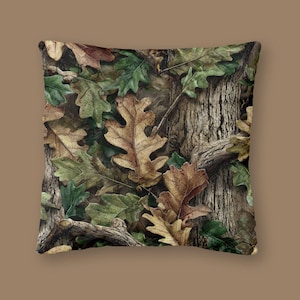 May include: Square cushion with a camouflage design of brown and tan oak leaves, green leaves, and tree bark. The pillow has a neutral background and is suitable for home decor.